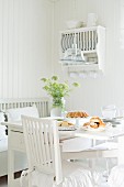 Table set for afternoon coffee with home-made pastries and jar of wildflowers in white, shabby-chic kitchen with old-fashioned plate rack on wall