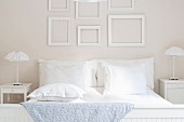 Weiß bezogenes Doppelbett, flankiert von Nachtkästchen mit Rüschenleuchten, an der Wand kassettenartig angeordnete, leere Bilderrahmen
