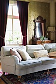 Pale couch with scatter cushions in front of red, gathered curtains on windows in traditional living room