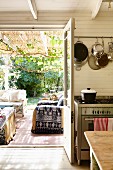 Blick von Küche durch offene Tür auf überdachte Terrasse mit gemütlichen Polstersofas, vor sommerlichem Garten