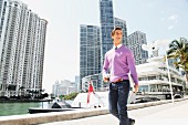 A young business man walking down the street holding a drink and a laptop with a harbour and office blocks in the background