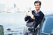 A young man wearing a jumper standing next to a car holding an iPad