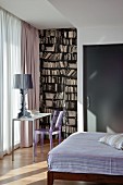 Designer lamp on small table and plastic chair in front of floor-length window curtains next to wallpaper mural with bookcase motif