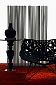 Schalensessel aus durchbrochenen Kunststoffelementen mit floralem Muster und ein Säulentisch mit Vase auf orangefarbenem Hochflorteppich