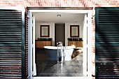 Blick durch offene Tür mit dunklen Türläden auf freistehende Badewanne mit Standarmatur in grossräumigem Bad