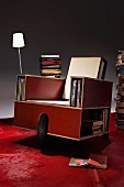 Multifunktionales rollbares Möbel aus beschichtetem Holz in Sesselform mit Regalöffnungen für Bücher und Tischleuchte, roter Boden