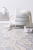 Weisser Polstersessel auf weißem Teppich mit Spitzenmotiv, im Hintergrund weiße Vase auf kubischem Bodentisch; edles feminines Ambiete