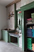Alte, grün lackierte Holzeinbauten in gefliester Wand mit Ketten-Deko; farbig sortierte, gestapelte Tücher in offenem Schrank