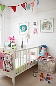 Cot, white wicker trunk, doll and toys in child's bedroom: children's clothing in picture frames on wall