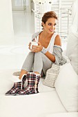 Junge Frau in bequemer Freizeitbekleidung, mit Winter-Hausschuhen, Tasse Tee und Wärmeflasche auf dem Sofa