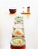 Five Korean side dishes in white bowls with soy sauce, salt and chilli powder in the background