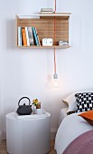 A breakfast tray on a white cylindrical bedside table with a pendant lamp with a naked light bulb and a small wall shelf above the bed