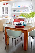 White shell chairs around wooden table in front of modern kitchen counter with bar stools and floating shelves