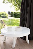Low, white table with glossy top and rustic legs next to glass wall with view into summery garden