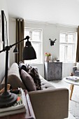 Retro table lamp next to pale couch, stylised hunting trophy on wall above vintage cabinet in background