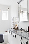 Stone washstand counter with white base cabinets and leather handles below mirrored cabinet and chandelier in white bathroom