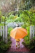 Kleines Mädchen mit orangefarbenem Regenschirm unter einem Rosenbogen im Garten