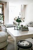 Vase of white peonies on white-painted table in simple living area with stool and stacked floor cushions