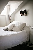 Schlichtes Bett mit Kissen und weisser Bettwäsche, unter Gaube mit Fenster, vor Wand mit aufgehängten Masken