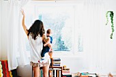 Young woman holding baby and opening bedroom curtains