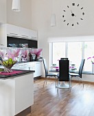 White, open-plan kitchen with magnolia-patterned splashbacks and dining area next to window below wall clock