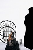 Ornamental structure of upturned wicker chair, wooden monkey on vase and profile of black tailors' dummy to one side