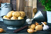 Kartoffeln in Metallschale, daneben Zwiebeln und Knoblauch auf altem Holztisch