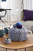 A knitted grey tea cosy decorated with colourful pompoms