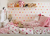 Love-heart pattern on bed linen and wallpaper in child's bedroom; collection of soft toys on wooden shelf and little blonde girl on metal bed