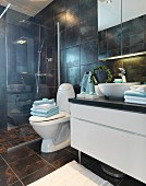 Washstand with countertop basin, pedestal toilet and floor-level shower with glass screen in dark-tiled bathroom