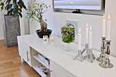 Lit, white candles in silver candlesticks on low, white sideboard in modern living room