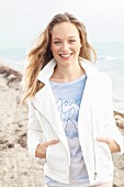 A young blonde woman on the beach wearing a shirt and a short, white jacket