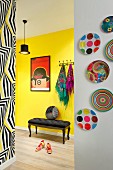 Framed red poster above antique, upholstered bench against yellow hallway wall, colourful plates on white wall and wall with patterned wallpaper