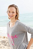 Junge, blonde Frau in grauem Pulli mit pinkfarbener Schrift am Meer