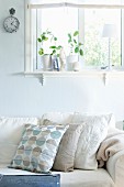 Sofa mit Kissen in verschiedenen Weissstönen vor pastellblau getönter Wand, unter Fenster