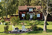 weiße Outdoor Stühle auf gekiester Fläche in sonnenbeschienenem Garten, im Hintergrund Landhaus mit rotbraun gestrichener Holzfassade