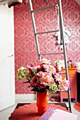 Blumenstrauss in orangefarbener Glasvase auf Spitzendeckchen, dahinter eine Metallleiter und pinkfarbenes Tapetenmuster