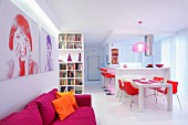 Modified portraits on wall above magenta sofa; white, open-plan kitchen and dining area with red chairs and accessories in background
