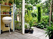 Kugelgrill und Regal mit Blumentöpfen auf Veranda mit Holzboden, vor sommerlichem Garten