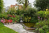 Flowering plants around small pond with stone figurine on edge next to small paved area