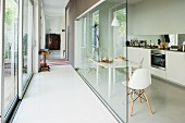 Schmaler Gang mit Glaswand, dahinter Essplatz mit Klassikerstuhl in Weiß, gegenüber moderne Küchenzeile
