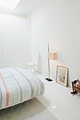 Bett mit gemusterter Tagesdecke, Stehleuchte und Einbauschrank in minimalistischem, weißem Schlafzimmer