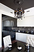 Chandelier hanging from ceiling panel in elegant kitchen-dining room with button-tufted chairs and white and glossy black cupboard fronts