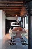 Elegant wooden counter and white swivel bar stools below pendant lamps and spotlights attached to power rail on wood-beamed ceiling in loft apartment with industrial character