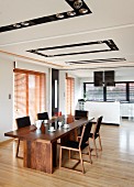 Elegant dining area with long wooden table and retro chairs in front of kitchen area in open-plan interior with recessed spotlights in ceiling panels