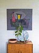 Sprig of leaves in vase and candlestick on wooden cabinet below artwork on wall