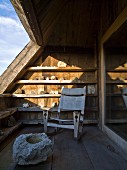 Rustikaler Sessel mit Segeltuchbezug auf Dachterrasse, vor sonnenbeschienenem Regal mit Steinsammlung, im Vordergrund Felsbrocken auf Holzdielen