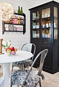 Dark display cabinet, round table, vintage chairs and shelves of ornaments on white wooden wall