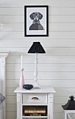 Still-life arrangement of pink candlestick, black bowl and table lamp with black lampshade on bedside table against white wooden wall