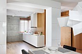 View across dining table in open-plan kitchen with white, minimalist counter and matching extractor hood against wooden wall